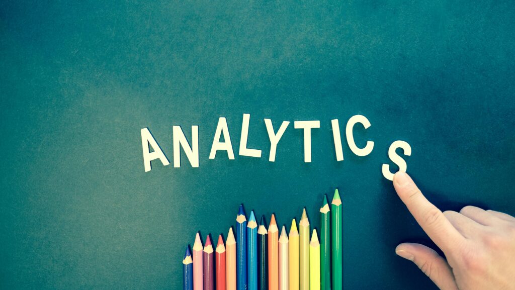 The word "Analytics" spelled out with letters above a row of colorful pencils, symbolizing data measurement and creative analysis in advertising performance tracking.
