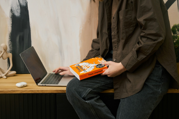 Marketer working on laptop while reviewing copywriting book for high-converting ads and landing pages.